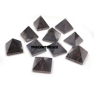 May include: A collection of ten dark gray pyramid-shaped stones. Each pyramid has a square base and a pointed top. The stones are arranged on a white surface. The text "THEGEMSSPLACE" is visible in the image.
