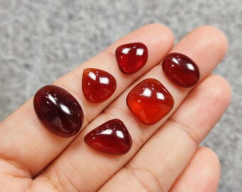 May include: Six red gemstone cabochons of various shapes and sizes, including oval, square, and teardrop, displayed on a hand.