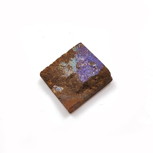 Op de afbeelding: Een vierkante, ruw getextureerde steen met een mix van bruine en paarse tinten. De steen heeft een natuurlijk, ruw uiterlijk, met een gedeelte van levendige paarse en blauwgroene kleuren. De algehele esthetiek suggereert een mineraal of edelsteen.
