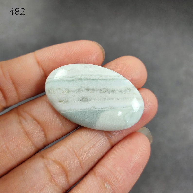 May include: A light green and white banded oval cabochon gemstone. The stone has a smooth, polished surface.
