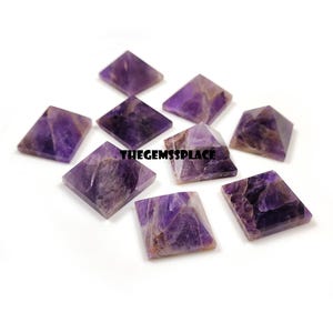May include: A collection of amethyst pyramid-shaped gemstones. The pyramids are a deep purple color with lighter purple and brown veining. The gemstones are arranged on a white surface. The text "THEGEMSSPLACE" is visible.