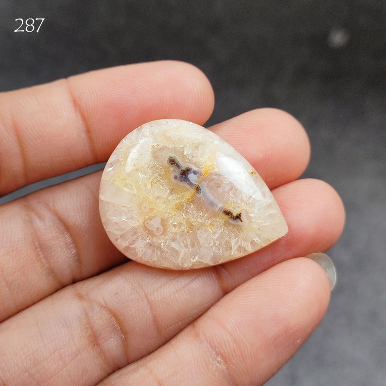 May include: A white agate cabochon with a purple and brown dendrite pattern. The cabochon is teardrop shaped and has a smooth, polished surface.
