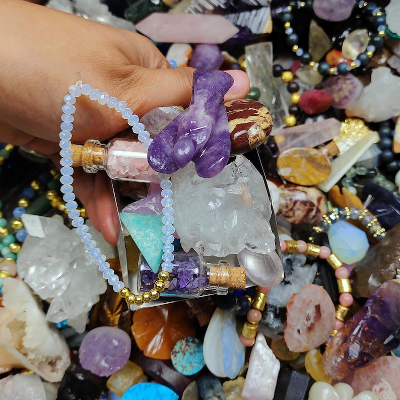 May include: A collection of crystals and gemstones, including a purple amethyst figurine, small glass bottles with cork stoppers, and a beaded necklace. The image showcases a variety of colors and textures, with a focus on natural stone jewelry and decorative items.