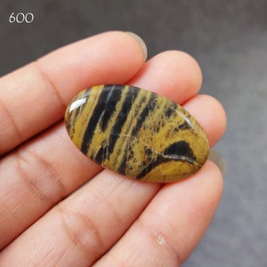 Op de afbeelding: Een close-up van een ovale cabochon edelsteen met gele en zwarte strepen. De steen heeft een gepolijste oppervlak en wordt in een hand gehouden.