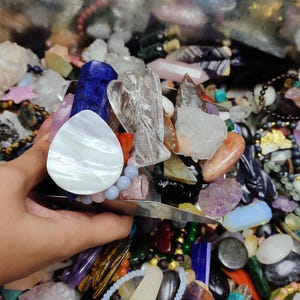 May include: A collection of colorful crystals and gemstones, including quartz, amethyst, and a carved angel figurine. The assortment features various shapes and sizes, with a pearlescent white stone and a blue crystal prominently displayed. Other items include beaded bracelets.