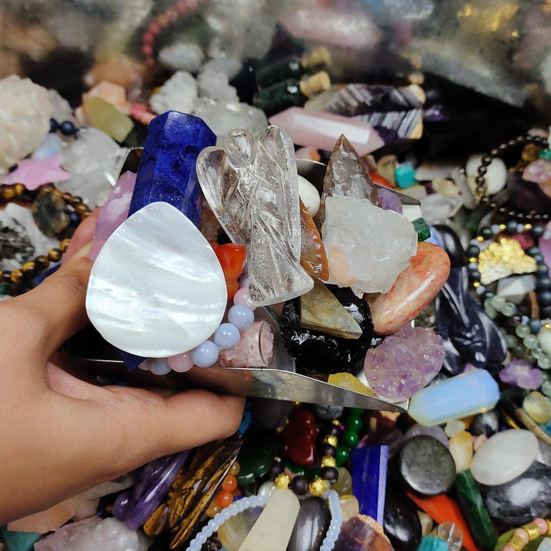 May include: A collection of colorful crystals and gemstones, including quartz, amethyst, and a carved angel figurine. The assortment features various shapes and sizes, with a pearlescent white stone and a blue crystal prominently displayed. Other items include beaded bracelets.