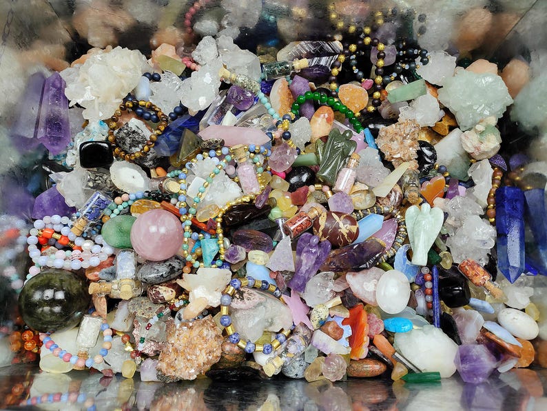 May include: A collection of various gemstones, crystals, and beads in a variety of colors. The assortment includes amethyst points, quartz clusters, and beaded bracelets. There are also small bottles and carved figurines.