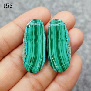 Puede incluir: Dos cabujones ovalados de malaquita verde y azul con un patr&oacute;n rayado. Las piedras se sostienen en una mano.