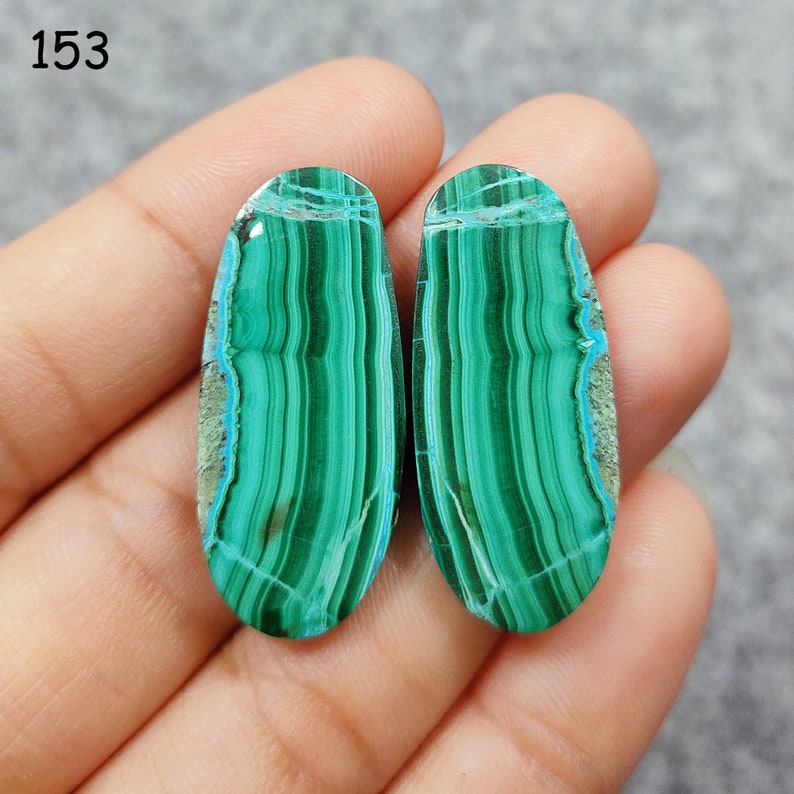Puede incluir: Dos cabujones ovalados de malaquita verde y azul con un patr&oacute;n rayado. Las piedras se sostienen en una mano.