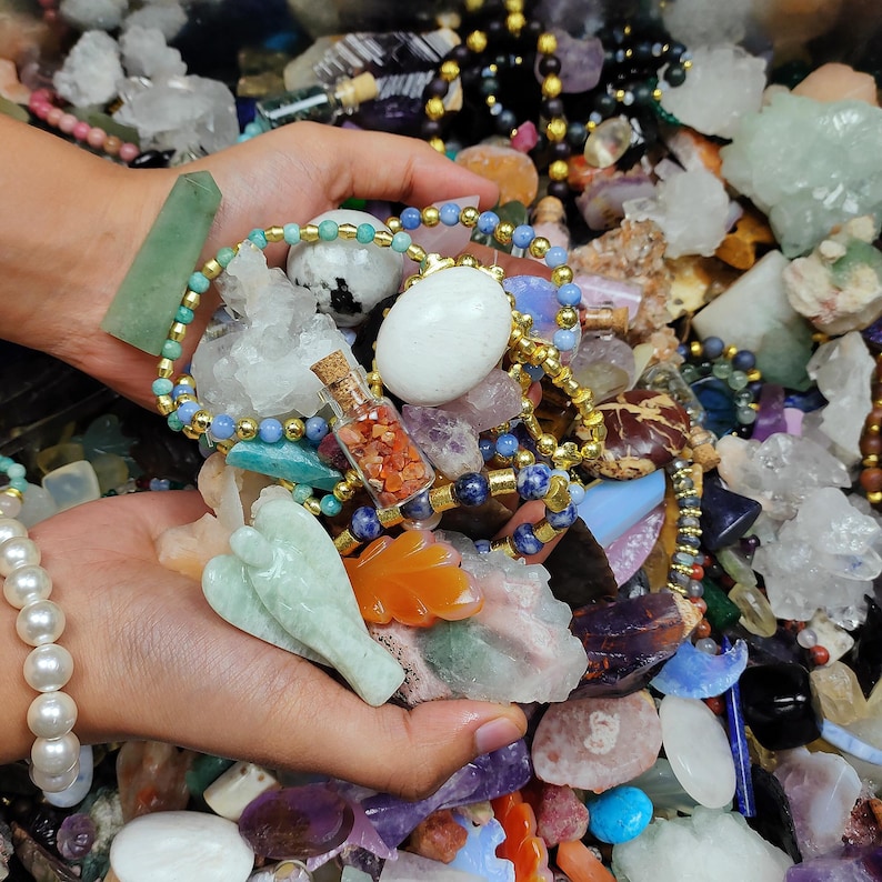 May include: A collection of gemstones and jewelry. The image features a variety of colorful crystals, beads, and small decorative objects. A beaded bracelet with gold accents and a small glass bottle with a cork stopper are visible.