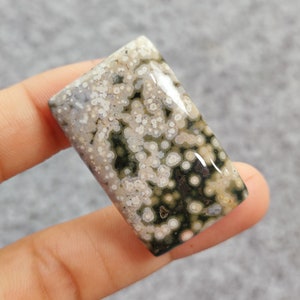 Op de afbeelding: Een close-up van een groene en witte oceaanjaspis cabochon. De steen heeft een uniek patroon van witte stippen en swirls op een groene achtergrond.