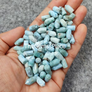 Puede incluir: Un montón grande de piedras preciosas azules y blancas en forma de pequeños cilindros puntiagudos. Las piedras están en una mano, y el texto "The GemssPlace" es visible en la imagen.