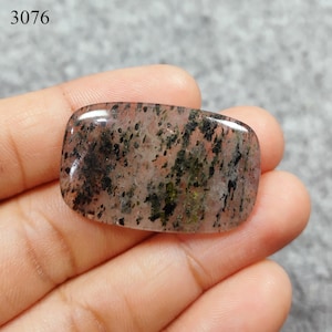 May include: A translucent, light pink, rectangular cabochon with black and green inclusions. The stone is approximately 2 inches long and 1 inch wide.