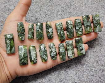 Puede incluir: Una colección de cabujones rectangulares de piedra preciosa de serafinita verde. Las piedras tienen una mezcla de tonos verdes oscuros y claros con inclusiones plateadas. Los cabujones se muestran en una mano.