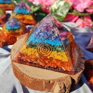 May include: A seven-chakra crystal pyramid with a copper wire spiral. The pyramid is made of clear resin with embedded crystals in the colors of the rainbow. The pyramid is sitting on a wooden base.