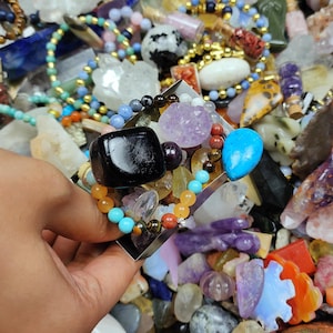 May include: A hand holding a silver-colored ring adorned with a large black stone, a turquoise teardrop, and various colorful beads. The background is filled with a variety of gemstones and crystals.