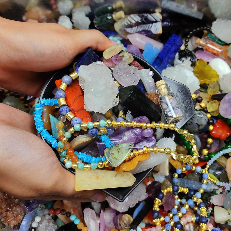 May include: A variety of colorful gemstones and crystals, including quartz, amethyst, and turquoise, are held in a black container. Gold beaded necklaces and small glass bottles are also visible. The image showcases a collection of natural stones and jewelry.