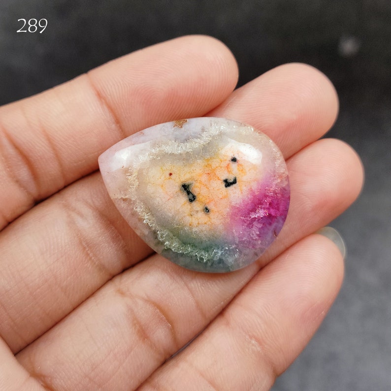 May include: A teardrop-shaped cabochon gemstone with a rainbow of colors, including pink, orange, green, and white. The stone has a unique, swirling pattern and is set in a clear, polished finish.