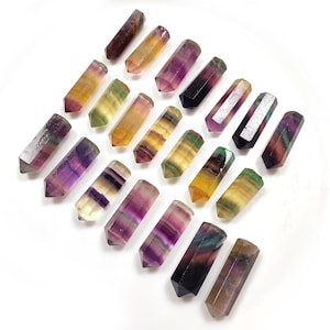 Puede incluir: Una colección de 18 puntas de cristal de fluorita de colores, cada una con un patrón único de rayas púrpuras, verdes, amarillas y blancas. Las puntas están dispuestas en un patrón escalonado sobre un fondo blanco.