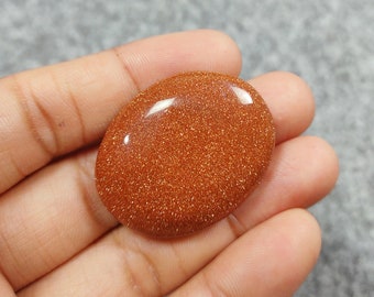 May include: A smooth, oval-shaped, orange-brown sunstone with a sparkly, glittery texture.