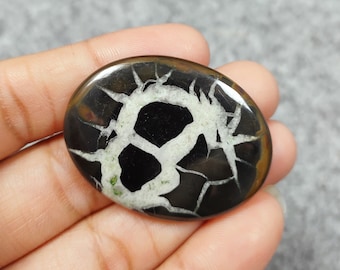 May include: An oval-shaped gemstone with a dendritic pattern in black and white. The gemstone has a brown rim and is set against a grey background.