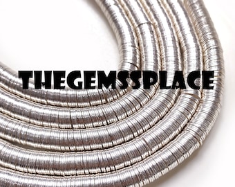 May include: Silver-toned metal beads, arranged in a circular pattern, forming a necklace or bracelet. The beads are flat and round, with a slightly textured surface. The image shows a close-up view of the beads.
