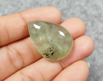 May include: A teardrop-shaped green gemstone cabochon with black inclusions. The stone is translucent and has a smooth, polished surface.