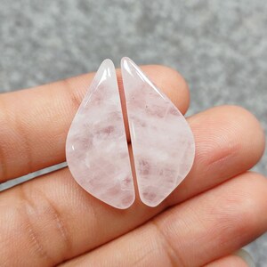 Op de afbeelding: Twee lichtroze rozenkwarts cabochons, elk in de vorm van een traan met een puntige punt, worden op een hand getoond.