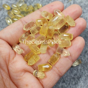 May include: A collection of yellow citrine gemstone points, also known as lemon quartz, in various sizes and shapes. TheGemsssPlace is printed on a hand holding the crystals.