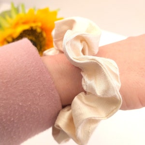 May include: A white scrunchie made of soft fabric, with a ruffled texture. The scrunchie is tied around a wrist.