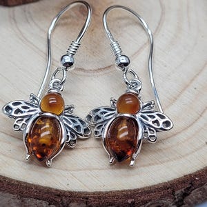 May include: Silver bee earrings featuring amber-coloured resin bodies and wings. The earrings have a hook closure. The earrings are displayed on a wooden surface.