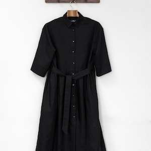 Può includere: Un abito camicia nero di lunghezza midi con colletto classico, abbottonatura frontale completa con bottoni metallici, maniche a gomito e una cintura in tessuto abbinata da annodare in vita. Il capo è rifinito con un orlo dritto.