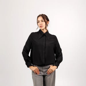 Op de afbeelding: Een zwart shirt met lange mouwen en een brede kraag. Het shirt is dichtgeknoopt en heeft manchetten met knopen. Het model draagt een grijze broek. De achtergrond is wit.