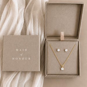 May include: A gold necklace with a single round clear stone pendant and matching stud earrings presented in a taupe gift box. The lid of the box is printed with the text MAID of HONOUR in white capital letters and a script font.