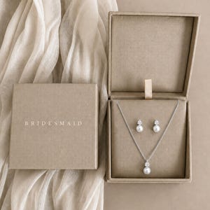 Könnte beinhalten: Ein Set aus einer silbernen Halskette und passenden Ohrsteckern, jeweils mit einer weißen Perle und einem klaren Stein. Der Schmuck liegt in einer offenen, quadratischen, beigefarbenen Schachtel, deren Deckel den weißen Schriftzug BRIDESMAID trägt.