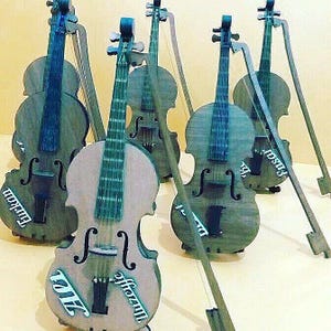 May include: A group of wooden violins with bows. The violins are in shades of grey and brown, with the strings and fingerboards in a contrasting colour. Some violins have text on them.