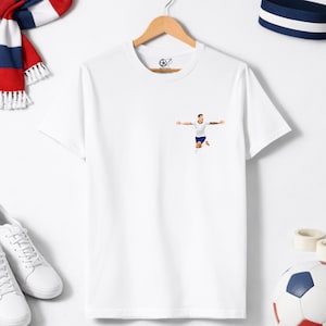 May include: White t-shirt with a small graphic of a soccer player celebrating a goal. The image also includes a soccer ball, white sneakers, a scarf with red, white, and blue stripes, and a blue headband.