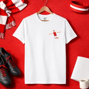 May include: A white t-shirt with a small graphic of a soccer player celebrating a goal. The shirt is displayed with a red and white scarf, a red and white headband, black soccer cleats with red laces, and other accessories on a red surface.
