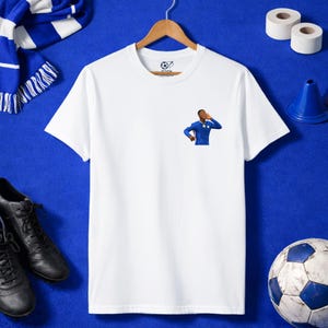 May include: White t-shirt with a small graphic of a soccer player in a blue uniform. The shirt is displayed on a wooden hanger, alongside a blue and white scarf, black shoes, a soccer ball, and training cones. The background is a solid blue.