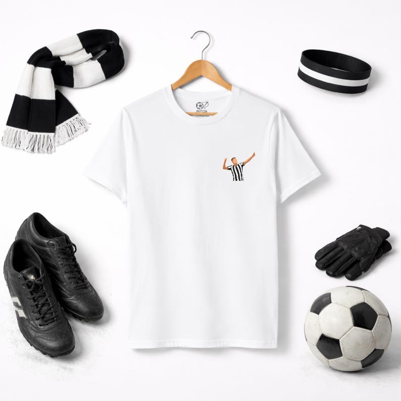 May include: A white t-shirt with a small graphic of a soccer player in a black and white striped jersey. The image also includes a black and white scarf, black soccer cleats, a black headband, black gloves, and a soccer ball.
