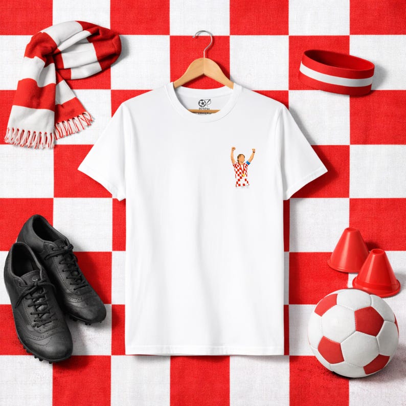 May include: White t-shirt with a small graphic of a soccer player celebrating, hanging on a wooden hanger. The image includes a red and white scarf, a soccer ball, and black soccer cleats. The background is a red and white checkered pattern.