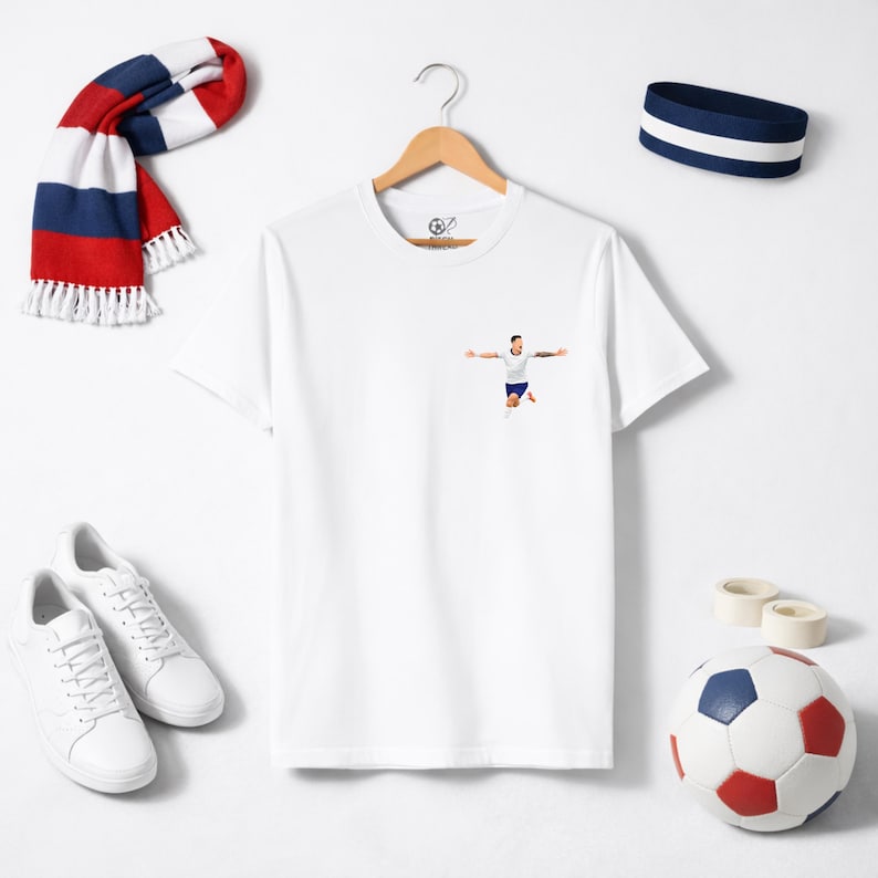 May include: White t-shirt with a small graphic of a soccer player celebrating a goal. The image also includes a soccer ball, white sneakers, a scarf with red, white, and blue stripes, and a blue headband.