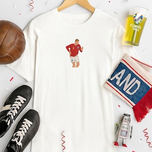 May include: White t-shirt with a graphic of a soccer player in a red jersey and white shorts. The image includes a soccer ball, black shoes, a glass of beer, a scarf, and a toy robot. The shirt is hanging on a wooden hanger.