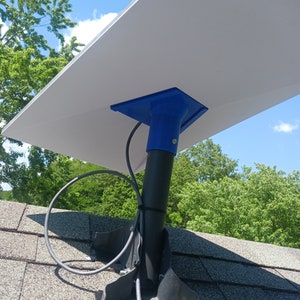 Puede incluir: Un panel solar blanco montado en un poste de metal negro con una base de plástico azul. El poste está unido a un soporte de metal negro en un techo de asfalto gris.