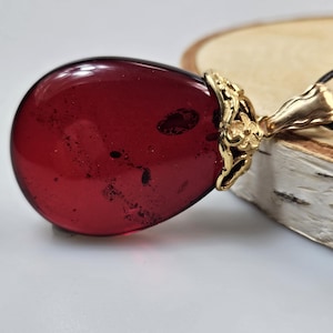 Puede incluir: Colgante de ámbar rojo oscuro en forma de lágrima con un engaste de filigrana dorada. El colgante cuelga de un cordón negro. El ámbar tiene inclusiones oscuras.