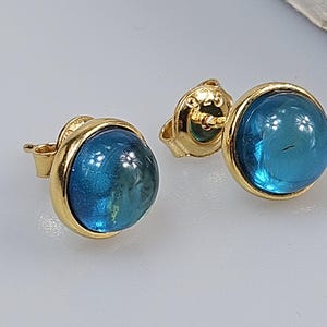 May include: A pair of gold-colored stud earrings. Each earring features a round, vibrant blue gemstone set within a gold bezel. The gemstones have a slight dome shape, and the earrings are displayed on a light-colored surface.