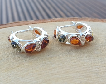 May include: A pair of silver hoop earrings with amber stones. The earrings are made of sterling silver and feature a variety of amber stones in different shades of brown and green.
