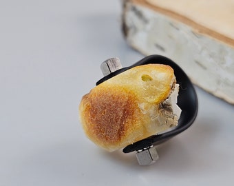 May include: A ring with a large, irregular piece of amber set in a black metal band. The amber is a warm yellow color with some white inclusions. The band is secured with two silver screws.