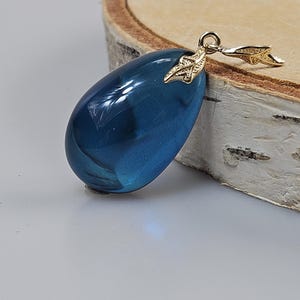May include: A deep blue, teardrop-shaped pendant with a glossy finish. It hangs from a gold-coloured bail with a leaf design. The jewellery piece is set against a light background and a wooden surface.