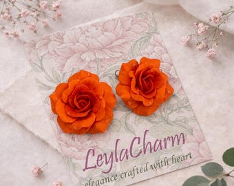 May include: Two vibrant orange rose-shaped stud earrings displayed on a rectangular card featuring a pale pink floral illustration and the text LeylaCharm elegance crafted with heart in a dark purple script font.